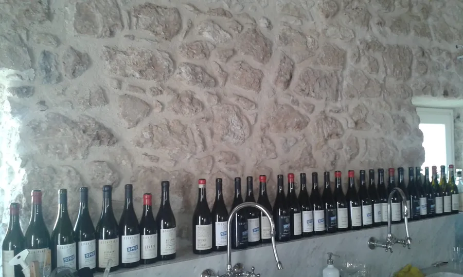 Una fila di bottiglie di vino su un ripiano di marmo, contro un muro di pietra