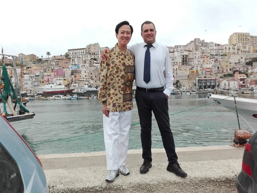 El chófer y una invitada en el muelle de un puerto, con las casas de colores detrás