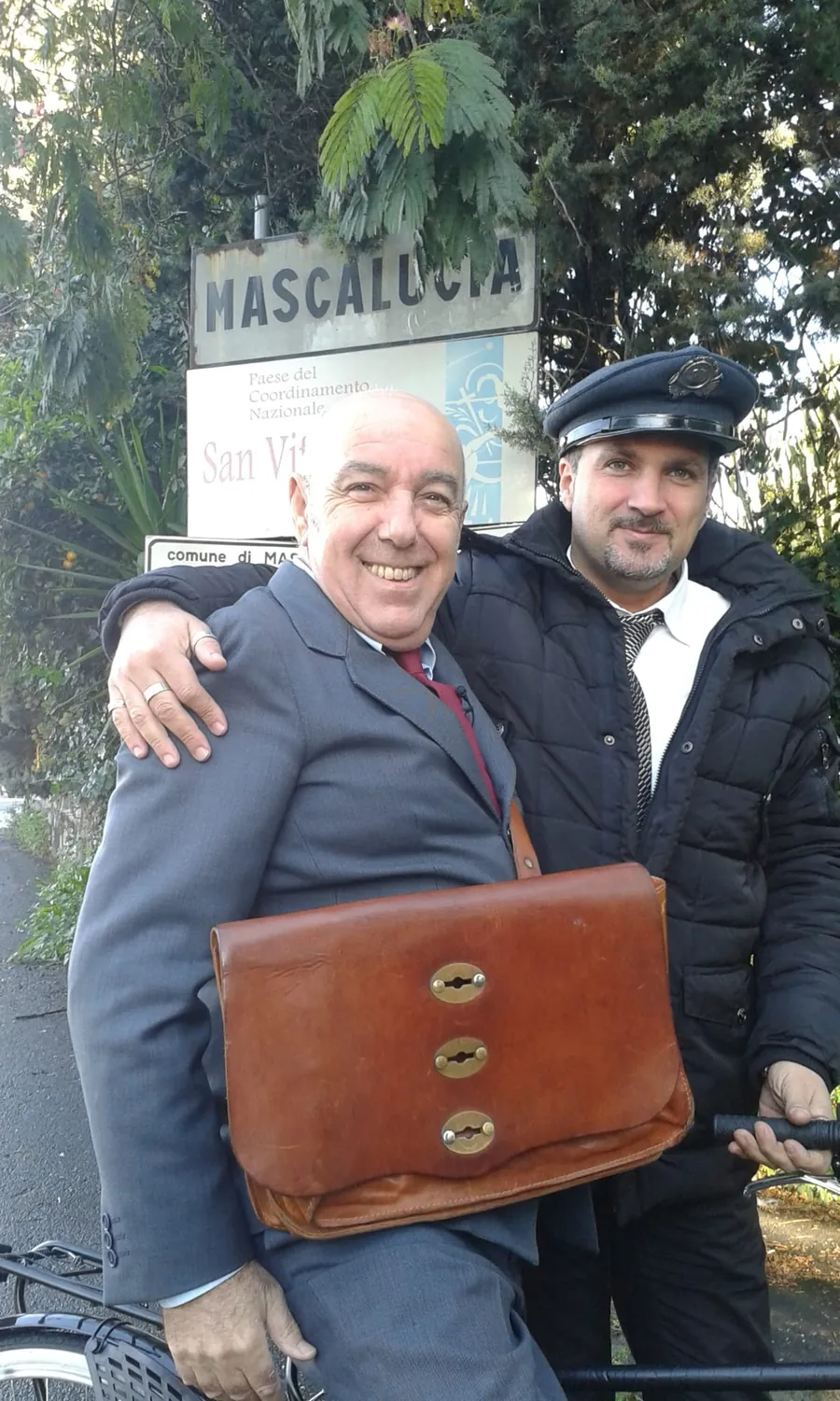 L'autista col berretto accanto a un ospite in abito con una borsa di cuoio, davanti al cartello di Mascalucia