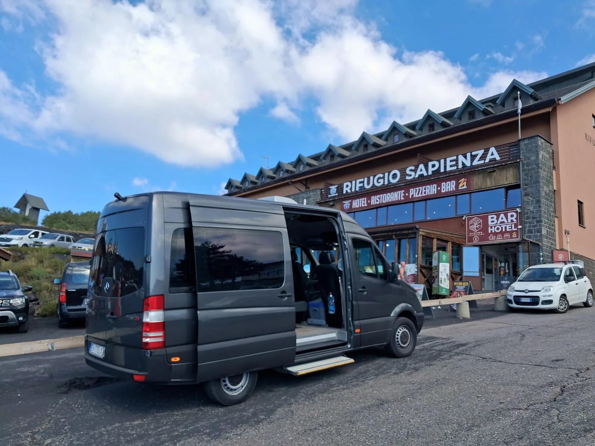 Il Mercedes-Benz Sprinter grigio scuro in un piazzale di montagna, con la porta laterale aperta e la pedana abbassata