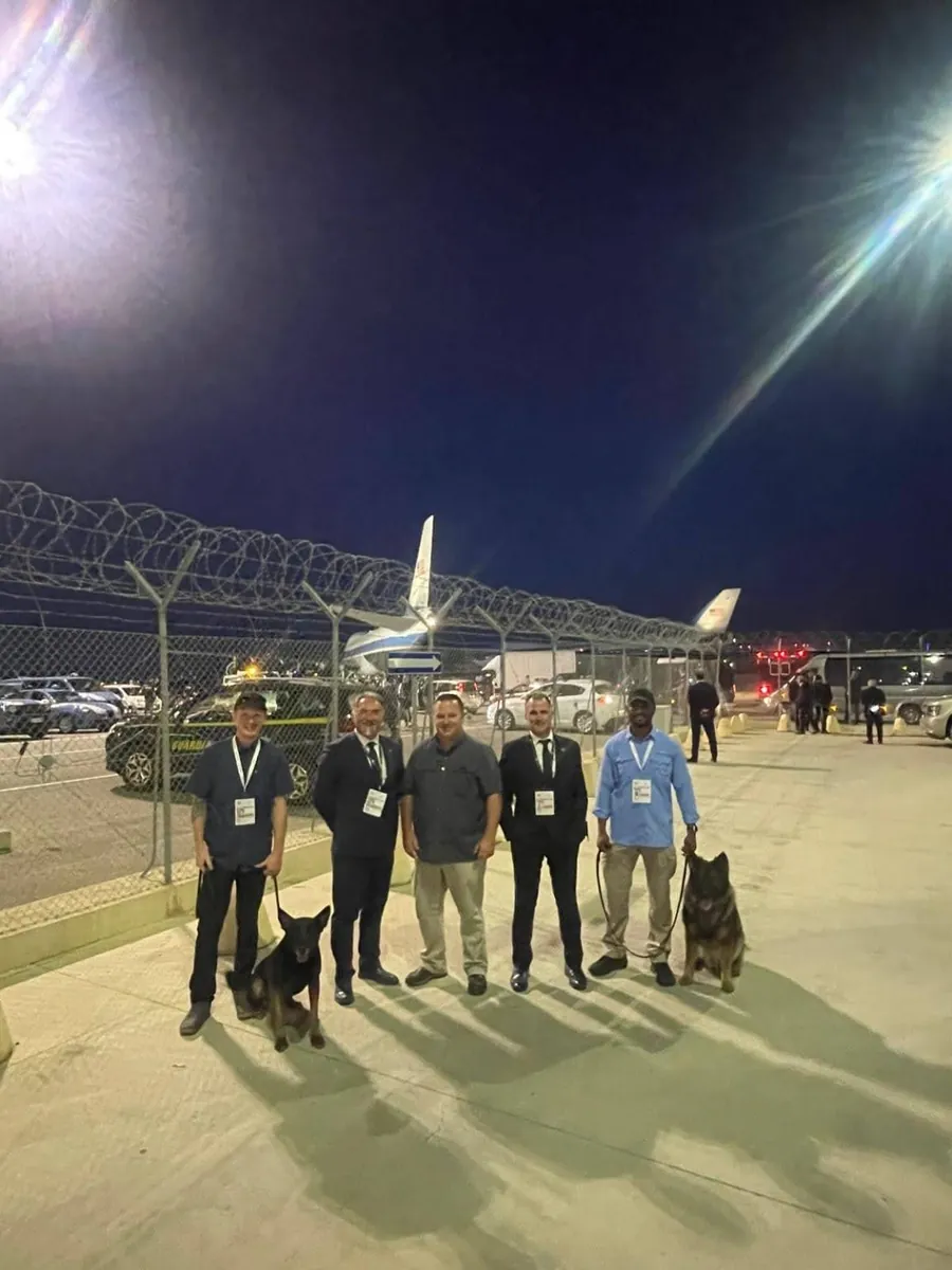 Di sera, in pista, cinque persone e due cani delle unità cinofile, davanti alla recinzione e a un aereo