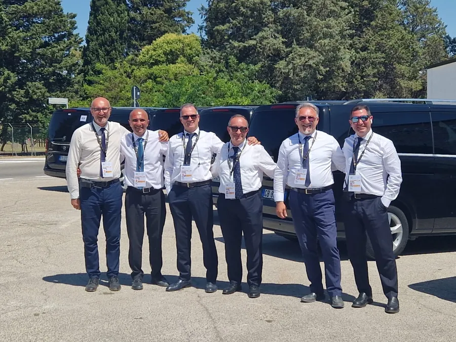 Sei autisti in camicia bianca e cravatta, con il pass del vertice, davanti alle auto nere