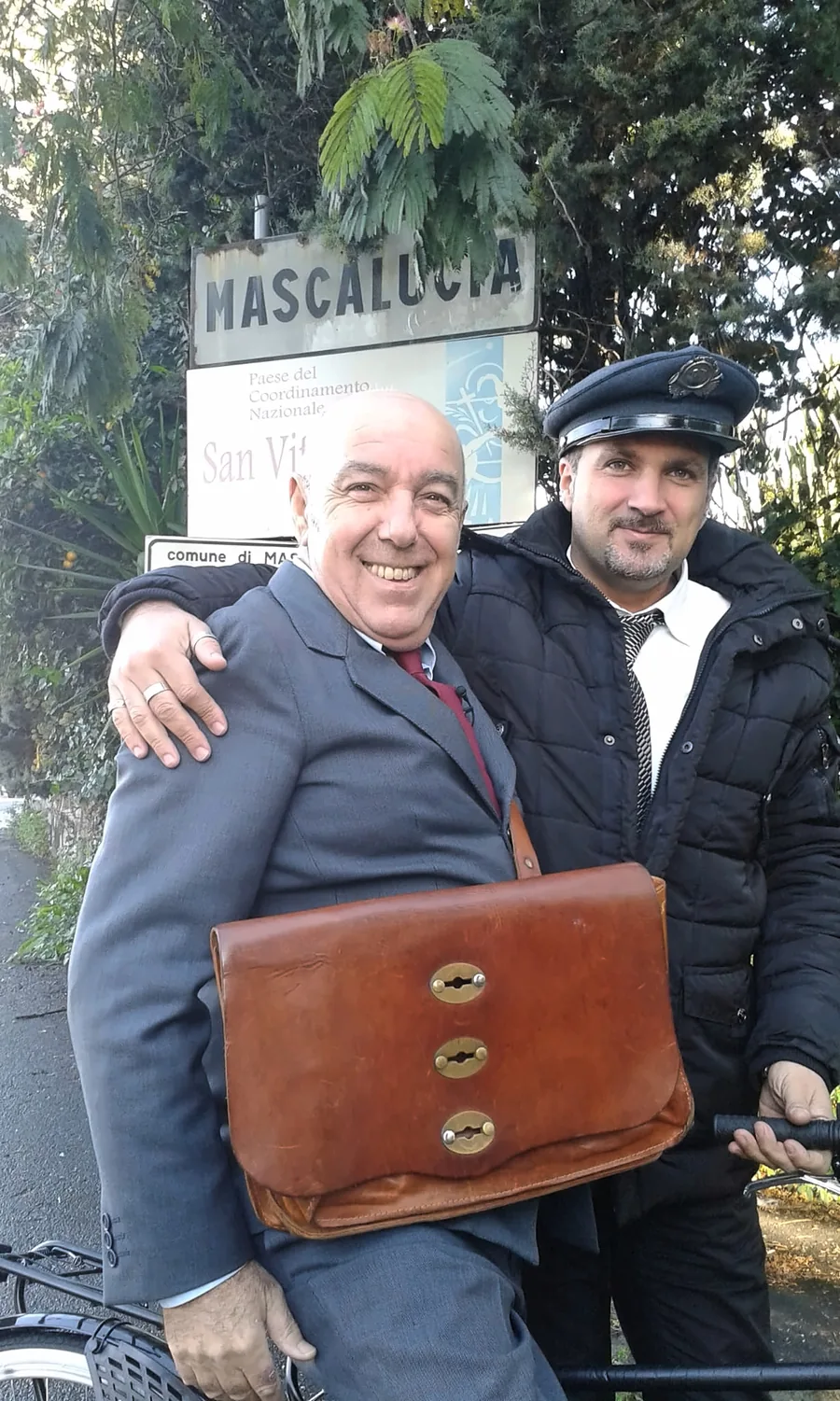 El chófer con gorra junto a un invitado de traje con un maletín de cuero, delante del cartel de Mascalucia
