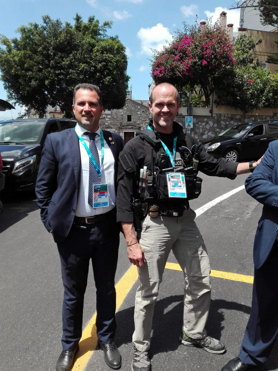 Un chófer con traje oscuro y el pase del G7, junto a un agente de uniforme, en Taormina